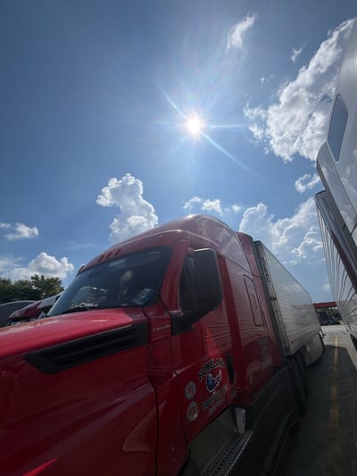 This is a low-angle, eye-level shot of the driver's side of a red semi-truck parked at a rest stop under a bright, sunny sky. The sun is visible in the upper left quadrant of the image, creating a lens flare effect. Puffy white clouds dot the bright blue sky. The truck features a white trailer, and another white truck is partially visible to the right. The truck has "HOMELAND TRANSPORT, INC." and "Buffalo Grove, IL" printed on its side, along with logos. A white disposable cup sits on the truck's running board. In the background, trees and a gas station canopy are visible. The scene is likely a truck stop or rest area, possibly in Lebanon, United States of America, given the presence of semi-trucks and the outdoor setting.