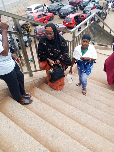 The image captures a scene on a pedestrian overpass in Immigration Quarters, Nigeria, with a focus on individuals descending a concrete staircase. The overpass has metal railings. In the foreground, a person wearing a light blue shirt and black trousers with black sandals is on the stairs. To their right, a woman in a black and gold patterned hijab and a red and gold patterned skirt is holding a black handbag. Further down the stairs, another woman in a green shirt and blue jeans is wearing a white shawl draped over her shoulders and a blue and white patterned fabric wrapped around her waist. She is holding a light pink handbag and wearing sandals. The background shows a street with various cars, including red, silver, and black vehicles, and a yellow auto-rickshaw. The overall environment appears to be a moderately busy urban setting.