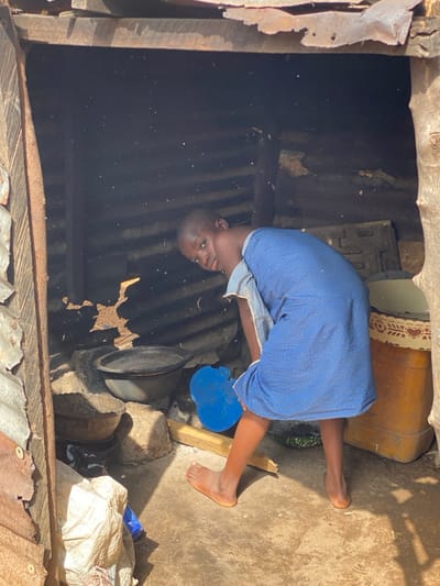 In Idiroko, Nigeria, an interior scene within a rudimentary structure reveals a young individual bent forward, looking directly at the camera. The person, barefoot and dressed in a blue patterned garment, has a light-colored cloth draped over their left shoulder. The structure features walls made of dark, corrugated metal sheets, some showing deterioration, supported by visible wooden beams. A makeshift cooking area is present on the left, comprising stones arranged to support two dark metal pots or bowls, one larger with a lid and one smaller. On the ground to the far left, a light-colored sack is visible. Near the individual's feet lies a blue, irregularly shaped plastic object and a wooden plank. To the right, a portion of a cream-colored cylindrical container with a decorative border is visible. The floor is an uneven mix of concrete and dirt. Strong sunlight enters from the opening, illuminating suspended airborne particles, likely dust or smoke, and creating sharp contrasts within the dark interior.