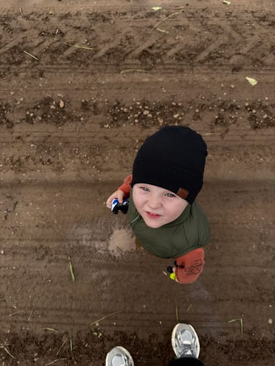 The image depicts an overhead view of a young child and the feet of an adult standing on a muddy, unpaved surface, located in Elovo, Russia.

The child, appearing to be a toddler or preschooler, occupies the mid-ground, looking directly upwards towards the camera with a distinct grimace or scowl. The child wears a black ribbed beanie with a small brown rectangular patch on the right side, an olive green puffer vest, and a long-sleeved orange or reddish shirt with a dark graphic design visible on the right sleeve. The child's left hand holds a small blue and white toy vehicle, and the right hand holds a small yellow object. The child's feet are positioned within a small, shallow puddle or wet patch of mud.

At the bottom of the frame, the lower legs and feet of an adult are visible, wearing dark trousers and grey and white athletic shoes.

The ground consists of dark brown, wet earth and mud, indicative of an outdoor, unpaved area. Numerous small white and grey pebbles are scattered across the surface. Distinct tire tracks run horizontally across the mud, suggesting a path or road. Scattered blades of green grass, some appearing broken or cut, are visible throughout the frame. Additionally, several pieces of light green leafy debris are present, particularly in the upper right portion of the image.
