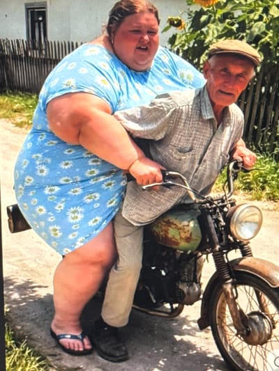 In Shcherbanka, Ukraine, an elderly man wearing a flat cap and a checkered shirt operates a rusty green motorcycle on an unpaved road. Behind him, a woman in a blue floral dress is seated. The background includes a wooden fence, a white building, and sunflowers.