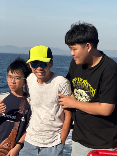 Three individuals are captured in a medium shot, posing outdoors against a backdrop of the ocean and distant hills under a clear sky. The person on the left, a younger male wearing a dark blue t-shirt and clear-rimmed glasses, has his left arm around the waist of the central individual. He is also holding a brown leather bag with a shoulder strap. The central figure, an adult male, is wearing a bright yellow baseball cap, sunglasses, and a light grey t-shirt. He has his right arm around the shoulders of the person on the right and is smiling. The individual on the right, a larger male, is wearing a black t-shirt with a graphic design and has his left arm around the waist of the central figure. The ocean occupies the background, with faint landmasses visible on the horizon. The location is identified as An Hải, Vietnam.