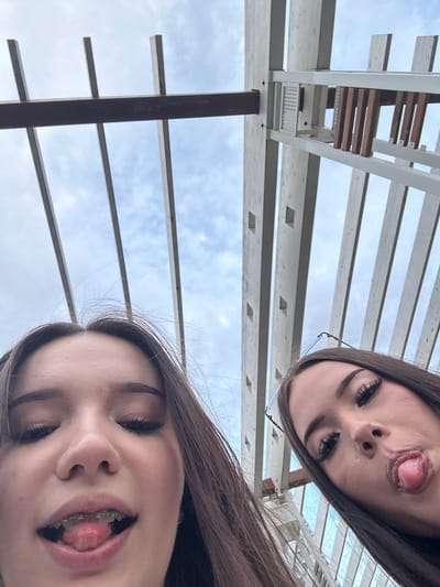Two young females are captured in a low-angle selfie, positioned in the foreground. The individual on the left is closer to the camera and has her mouth open, revealing braces on her teeth, and her tongue is protruding. The individual on the right is slightly further back, also with her mouth open and tongue sticking out. She is wearing glasses that rest on her nose. Both subjects have dark hair and appear to be outdoors.
The background consists of a structure made of light-colored beams, possibly a pergola or similar architectural element, against a sky with visible clouds. The perspective suggests the photo was taken from below, looking upwards at the subjects and the sky. The scene takes place outdoors in an urban environment, likely Naberezhnye Chelny, Russia, given the context.