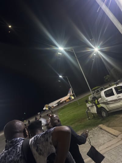 The image captures a scene at night, likely in Omoecheigbo, Nigeria. Several people are visible in the foreground. In the background, an airplane is parked on the tarmac next to what appears to be a white truck. The area is illuminated by tall light posts. A purse is visible in the foreground. The sky is dark.