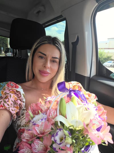 A blonde woman with styled hair, makeup, and a floral print dress sits in the passenger seat of a vehicle. She is holding a bouquet of flowers in a basket tied with a purple ribbon. The woman's face is partially obscured by the bouquet. The interior of the car is visible, including the headrest of the driver's seat and part of the window. Outside the window, a street scene with buildings and trees can be seen under a cloudy sky. The location is Pazardzhik, Bulgaria.