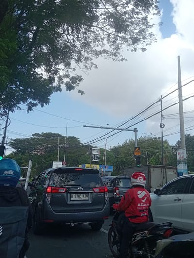 The image depicts a street scene in Surabaya, East Java, with heavy traffic. In the foreground, a person wearing a red jacket and helmet, likely a delivery rider, is seated on a motorcycle. The red jacket has "ACI" and a heart logo prominently displayed. Behind the motorcycle, a dark-colored Toyota Innova is visible, with a license plate that reads "B 2701 KIU". Several other vehicles are present, indicating a traffic jam. Overhead, power lines and traffic lights crisscross the sky, which is partly cloudy. Lush green trees line the background, and various advertisements and signs are posted along the roadside, including one that reads "AREA DILARANG CCTV". The overall atmosphere suggests a busy urban environment during daylight hours.