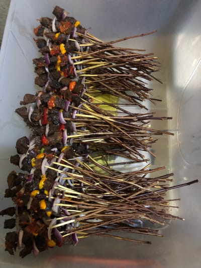 A vertical, eye-level, medium shot captures a close-up of many beef skewers, each prepared with pieces of marinated beef, red onion slices, and small pieces of red and yellow bell peppers, all threaded onto thin, brown wooden skewers. The skewers are arranged in a large, clear plastic container, with the meat portions concentrated on the left side of the frame and the wooden skewers extending to the right. The beef appears dark brown and slightly charred, suggesting it has been grilled or is ready for grilling. The onions are a vibrant purple-white, and the peppers add pops of color. The container is a light gray, translucent plastic, and a sliver of a yellow surface is visible beneath some of the skewers on the left. The background is a blurred, light gray, and a hint of the upper edge of the container is visible. The scene appears to be set indoors, likely in a kitchen or food preparation area, possibly in Kabong, Nigeria, given the context of the prompt. There are no people visible in the image. The lighting is even, highlighting the textures and colors of the food.