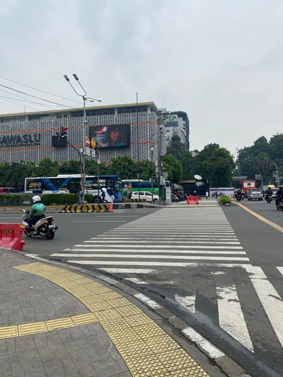This image captures an urban scene in Central Jakarta, Indonesia, likely during the daytime given the overcast sky. The foreground features a pedestrian crosswalk with prominent white stripes on the asphalt road. Adjacent to the crosswalk is a tactile paving surface, indicated by the textured yellow tiles, designed to assist visually impaired individuals. A motorcycle with a rider wearing a light green helmet and jacket is visible on the right side of the frame, positioned near the edge of the road.
Further into the scene, several vehicles are present, including buses, one of which is labeled "ELECTRIC BUS" and "Transjakarta". The buses are stopped or moving slowly, suggesting traffic or a bus stop. In the background, a large building with a distinctive white, patterned facade dominates the skyline. A large digital billboard on this building displays an image of a person's face. Trees line the street, providing greenery amidst the urban environment. Traffic lights are also visible, indicating an intersection. The overall impression is one of a busy street in a city, with a mix of vehicles, pedestrians, and urban infrastructure. The weather appears to be cloudy, contributing to the diffused lighting in the image.