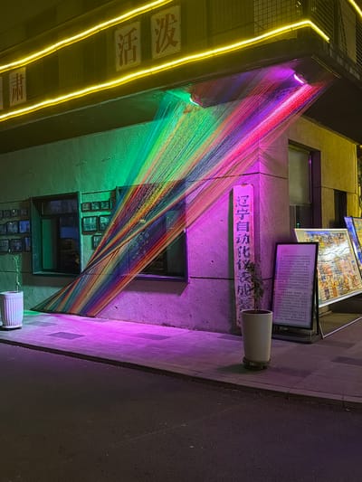 Here's a description of the image:
The image depicts a nighttime urban scene, likely in Linghe, China, given the presence of Chinese characters and signage. A building facade is illuminated with a vibrant, multicolored art installation consisting of numerous strands stretched diagonally across windows and other architectural features. The colors transition from green to purple.
Yellow neon lights are visible on the upper left. Various signs are mounted on the building facade, including one with an advertisement. A small plant in a cylindrical pot is mounted to the wall as well. The sidewalk is visible on the right and a road is visible in the right background.