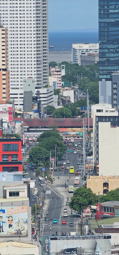 This is an elevated, eye-level, vertical, eye-level, long shot photograph capturing a bustling street scene in Makati, Philippines. The image is dominated by high-rise buildings, a busy road with numerous vehicles, and lush green trees.

On the left side of the frame, a tall, white residential building with numerous windows stretches towards the sky. Adjacent to it, a narrower, beige building with distinct horizontal lines of windows stands out. Further down, a vibrant red, multi-story building with a grid-like facade of windows is prominent. Below this red building, a billboard features cartoon characters and text.

The road in the center of the image is filled with traffic, including cars, buses, motorcycles, and tricycles (motorcycle with sidecar). Vehicles are moving in both directions, indicating a flow of urban transportation. Traffic lights and street signs are visible, suggesting an organized urban infrastructure. Tall utility poles with a dense network of wires crisscross the scene, a common feature in Philippine cities.

Green trees, some with full canopies, are interspersed throughout the urban landscape, offering a contrast to the concrete and glass structures. Some trees are planted along the roadside, while others are part of larger green spaces.

In the background, more buildings of varying heights and architectural styles can be seen, including a modern glass-fronted skyscraper on the right. The sky is a clear, light blue, suggesting a sunny day. In the far distance, a body of water is visible, possibly Manila Bay, with a ship on the horizon, hinting at the city's proximity to the coast. The scene conveys a sense of active urban life and the typical density of a major metropolitan area.