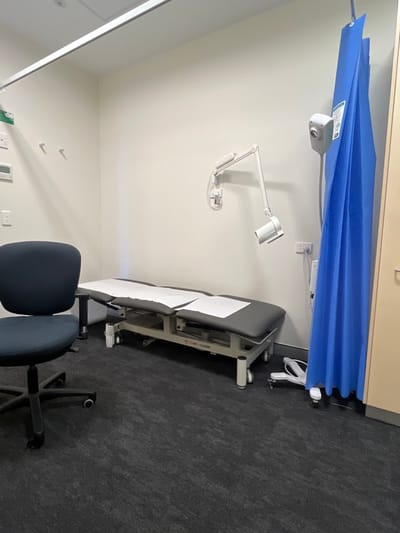 An examination room features a gray examination table with paper covering, placed against a gray wall. An adjustable examination light is mounted on the wall above the table. A blue privacy curtain hangs from the ceiling near the top of the image. In the foreground, a blue office chair sits on a gray floor. Wall-mounted electrical outlets and light switches are visible in the lower left.