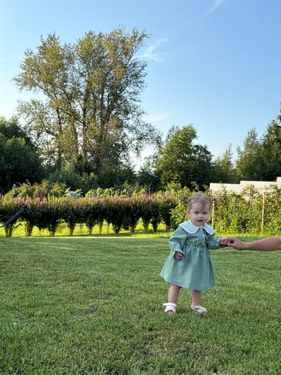 In the city of Garevaya, Russia, a young girl stands in a well-manicured grassy yard, holding the hand of an unseen adult. The girl, with light brown hair in two pigtails, wears a green dress with a white collar and white sandals. She is looking towards the camera with a neutral expression. The background features a dense line of bushes with pink flowers, mature trees, and a white fence. The sky is clear and blue, suggesting a sunny day. The scene is set in an outdoor, natural environment, likely a garden or park, with a focus on the child and the surrounding greenery.