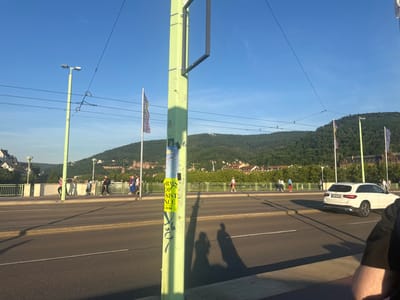The image shows a street scene in Heidelberg, Germany, likely taken on the Theodor Heuss Bridge looking east. The bridge spans the Neckar River, with views of the Old Town and the Heidelberg Castle on the hillside. A light green lamppost stands prominently in the foreground, covered with stickers and casting shadows. Pedestrians are walking on the pedestrian walkway of the bridge, and a white Mercedes-Benz SUV is driving on the road. Flags are seen in the background near the castle. The sky is clear and blue.