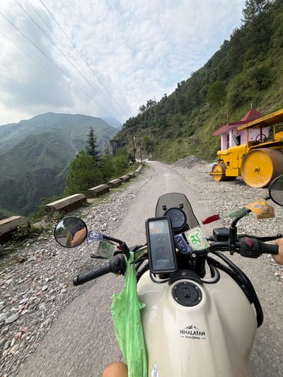 This media file is a first-person perspective video recorded from a motorcycle on a winding road in Dharamshala, India. The scene is mountainous and rural, with lush green hills and a cloudy sky. 

The primary subjects are the motorcycle and its rider, though only parts of the rider are visible, such as a hand on the handlebar and a leg. The motorcycle is a cream-colored Royal Enfield Himalayan, identifiable by the "HIMALAYAN ROYAL ENFIELD" decal on the fuel tank. Attached to the handlebars are prayer flags, common in this region, adding a cultural detail to the scene. A phone mount with a device is also attached to the handlebars.

In the background, a yellow road roller is parked on the side of the road. The road itself is paved with gravel and asphalt, and a concrete barrier lines the edge of the mountain, suggesting a steep drop. 

The time of day appears to be daytime, under overcast skies. The overall mood is one of travel and exploration through a scenic, mountainous landscape. There are no clear indications of specific events, but the act of riding suggests a journey.