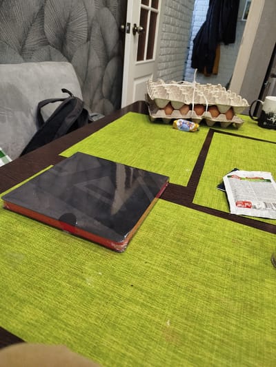 This image depicts a table set with a lime green placemat. On the placemat, there is a stack of black and red notebooks, an egg carton containing brown eggs, a small candy wrapper, and a packet of spice. In the background, a gray couch and a backpack are visible, along with a doorway and a wall with a textured pattern. The scene appears to be indoors, possibly in a residential setting, and suggests a casual environment. The presence of the placemat and the items on the table hint at a meal or a work-related activity.