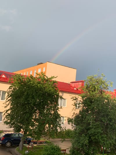The image depicts a scene in the city of Kapyĺ, Belarus, following a rain event, as evidenced by a rainbow arching across the sky above apartment buildings. The buildings have light-colored facades and red tile roofs. Lush green trees with dense foliage are prominent in the foreground, partially obscuring the lower floors of the buildings. A dark blue station wagon is parked on the paved area in front of the building, near a small playground area with benches and a sandbox. The sky is overcast with a gradient of gray, indicating residual clouds from the rain. The overall atmosphere is calm and serene. There are no people visible in the image.