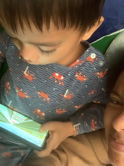 The media file shows a close-up, overhead view of two people, likely a child and an adult, in a relaxed setting. The child is wearing a dark blue pajama set with a pattern of orange crabs and white wavy lines. A white button is visible on the pajama top. The child's head and shoulder are in the foreground, with dark hair and closed eyes, suggesting they might be sleeping or resting.
The adult's face is partially visible in the upper right corner, with their lips close to the child's head. They are wearing a tan-colored garment. One of the adult's hands is visible holding a smartphone, which is displaying a bright, colorful screen with a scene that appears to be from a video game or animation. The screen shows vertical lines and what looks like a hallway or interior scene.
The background is composed of various soft textures, including a dark blue surface, a green and white striped fabric, and a gray, fuzzy material, possibly bedding or a blanket. The lighting appears to be indoor and soft, with no strong shadows, suggesting it could be daytime or evening. There is no visible text in the image. The overall impression is one of intimacy and comfort, with the adult sharing a quiet moment with the child, possibly while the child is resting or engaged with the phone. The location context provided is Sungai Meriam, Indonesia.