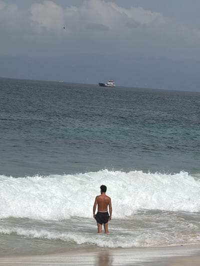 In the city of Manggis, Indonesia, a bare-chested individual wearing black swim trunks stands facing the ocean, with the lower half of their body submerged in the water. The person is positioned in the foreground, with a large wave breaking behind them, creating a foamy white crest and spray. The water in the foreground is relatively calm, with smaller ripples. In the mid-ground, the vast expanse of the ocean stretches towards the horizon, appearing dark and deep blue. Further out, a white vessel with red accents, possibly a ferry or a smaller cargo ship, is visible. In the background, a hazy mountain range is discernible against a cloudy sky. The clouds are a mix of white and gray, with a small dark bird flying high above the water. The overall scene depicts a serene beach environment with a solitary figure enjoying the ocean.