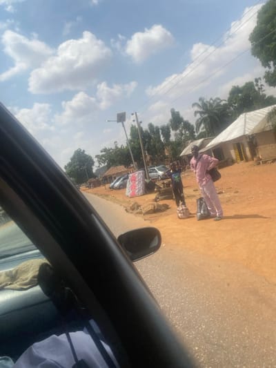 The scene depicts a daytime roadside in Tataura, Nigeria, viewed from within a vehicle. On the right side of an asphalt road, two individuals stand on a reddish-brown dirt shoulder. An adult male, wearing a light-patterned long-sleeved shirt and trousers, has a dark shoulder bag and stands beside a dark piece of luggage. To his left is a child or young male, clad in a dark t-shirt with a green graphic and dark trousers, standing next to a light-colored patterned bag. A large, upright white and red patterned mattress is visible on the dirt shoulder, along with several rocks and a discarded tire. In the background, modest buildings with corrugated metal roofs, parked vehicles, various trees including palm trees, and utility poles with overhead power lines are visible under a blue sky with scattered white cumulus clouds. The interior of the camera-operating vehicle, including the A-pillar and side mirror, partially frames the left side of the image.