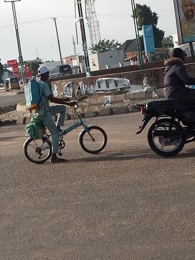 In Pantame, Nigeria, a man wearing a light blue outfit and a white cap cycles a blue foldable bicycle. He carries a large blue sprayer on his back and a green bag strapped to the rear of the bicycle. The man's right foot is resting on the pedal while his left foot is on the ground, indicating he is either starting or stopping. He is on a paved road with a sidewalk and roadside clutter in the background, including posters, buildings, and utility poles. To the right, a person on a black motorcycle is partially visible, also traveling on the road. The scene suggests an outdoor urban environment with typical street activity.