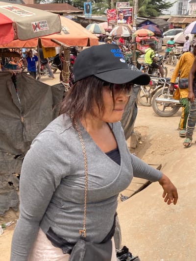 A forensic examination of the image reveals an outdoor market scene in Ura Nta Umuarandu, Nigeria.
The primary subject is a woman positioned in the foreground, facing right and slightly away from the camera. She wears a black baseball cap with "GLOBAL REIGN" in blue text, dark sunglasses, a grey long-sleeved ribbed wrap-style top over a black undershirt, and a light-colored skirt or lower garment. A black crossbody bag with a gold chain strap is visible across her torso. Her dark hair falls to her shoulders, and her left hand is partially visible, appearing to be in motion.
The background depicts a bustling market environment with a predominantly dirt ground and uneven terrain. Numerous individuals are scattered throughout the scene, some walking or standing near market stalls. Multiple motorcycles are parked or being operated. Market infrastructure includes several awnings and umbrellas, one prominently displaying "POWER HORSE ENERGY DRINK" branding. Dark tarpaulin sheets form makeshift shelters or stalls. In the midground and background, various buildings are visible, alongside multiple large posters and banners affixed to poles, some appearing to be political campaign advertisements (e.g., "PRINCE," "EMMANUEL ANYANWU"). Overhead utility lines are present. The lighting suggests a bright, sunny daytime setting.