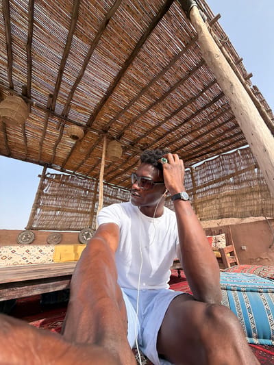 A man with dark skin is sitting on cushions under a thatched roof structure. He is wearing a white t-shirt, light blue shorts, sunglasses, and earbuds. His right hand is touching his hair. A watch is visible on his left wrist. The background shows a desert landscape with a clear sky. The scene appears to be a lounge area in a resort or camp in Agafay, Morocco, indicated by the traditional Moroccan decor and desert environment.