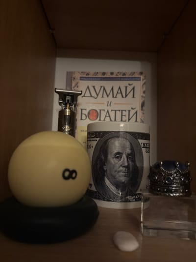 In a dimly lit room, on a wooden shelf, a collection of objects is arranged. In the foreground, a yellow billiard ball with the number "8" in black is prominently displayed, resting on a black base. To its right, a white mug features a portrait of Benjamin Franklin, a prominent figure from US currency, surrounded by the imagery of a hundred-dollar bill. Behind the mug, a book with Russian text, including the words "ДУМАЙ" (Think) and "БОГАТЕЙ" (Rich), stands upright. To the left of the billiard ball, a metallic hair trimmer with a silver and gold finish is positioned. Further to the right, a decorative silver ring with blue stones, shaped like a crown, sits on a clear glass object. The background of the scene is the dark interior of a shelf. The overall impression is one of personal belongings arranged on a display shelf.
