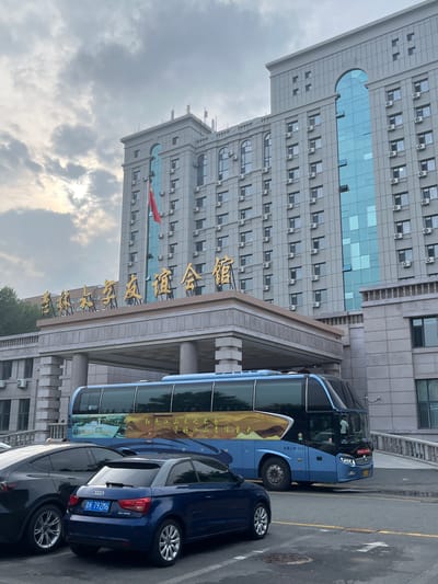 The image captures a wide shot of a large, modern building in Changchun City, China. The building is gray and features numerous windows and air conditioning units. Above the entrance, large golden Chinese characters are prominently displayed. A bright blue bus with a scenic mural on its side is parked in front of the building's entrance, partially obscuring the lower portion of the structure. In the foreground, two cars are parked: a dark-colored sedan on the left and a smaller blue Audi on the right. The sky above is cloudy, with sunlight filtering through the clouds. The overall scene appears to be an exterior shot of a hotel or a large public building.