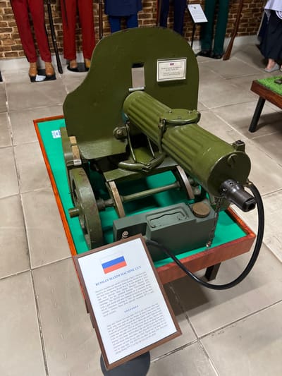 A green Russian Maxim Machine Gun, model 1910, sits on a wooden stand. A sign with the Russian flag indicates the gun's name. The sign describes it as a heavy machine gun used by the Imperial Russian Army during World War I and the Red Army during World War II. It was adopted in 1910 and chambered for the 7.62x54mmR cartridge. The M1910 was mounted on a wheeled mount with a gun shield. It was replaced in Soviet service in 1941.