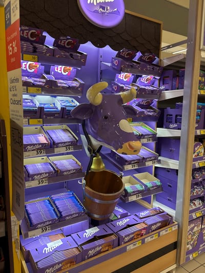 The image depicts a dedicated Milka chocolate display inside a supermarket, likely a Tesco store, in the Capital City of Prague, Czechia, identifiable by the "Kč" currency and "Clubcard" loyalty program signage. The central feature is a large, three-dimensional purple Milka cow head with yellow horns, white patches, and a light brown snout, wearing a brass bell suspended from a black strap and a wooden bucket below its chin. The display is predominantly purple, showcasing numerous Milka products on multiple shelves. Upper shelves feature "Milka LEO" bars, priced at 16 Kč and 23 Kč. Middle shelves present various Milka chocolate bars, many marked at 39.90 Kč. The bottom shelves display larger Milka chocolate boxes, including "Milka Max" variations, priced at 79.90 Kč. A prominent yellow sign on the left indicates a "15% sleva" (15% discount) and savings up to 15% with a Clubcard application. Overhead, a circular, illuminated "Milka" sign with the brand's iconic cow logo is visible. The shelving unit is illuminated from within, highlighting the products. No individuals are present in the scene.