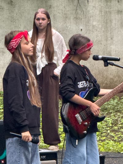 The image shows three young females on an outdoor stage with a concrete wall in the background and foliage to the right. A girl in the foreground on the left wears a red bandana and a black sweatshirt, holding a microphone in her right hand. Her left hand is on a microphone stand. To her right, another girl, also wearing a red bandana and a black sweatshirt, plays a red electric guitar. A third girl stands behind them, wearing a white top and brown pants. The scene appears to be a performance in the city of Dmitrov, Russia.