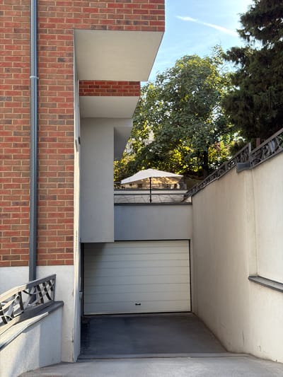 The image shows an exterior view of a building's facade and an adjacent garage. The building features a brick base with a light-colored stucco upper section. A large, light-colored garage door dominates the right side of the image. Above the garage door, a section of the building has a textured, light-colored wall. The scene is bathed in sunlight, suggesting it is daytime. Lush green trees with visible foliage are present in the upper left portion of the frame, indicating an outdoor setting with vegetation. A metal railing or decorative element is visible along the top edge of the building. The overall impression is one of a residential or commercial property, possibly in a suburban or urban environment. There is no visible text in the image.