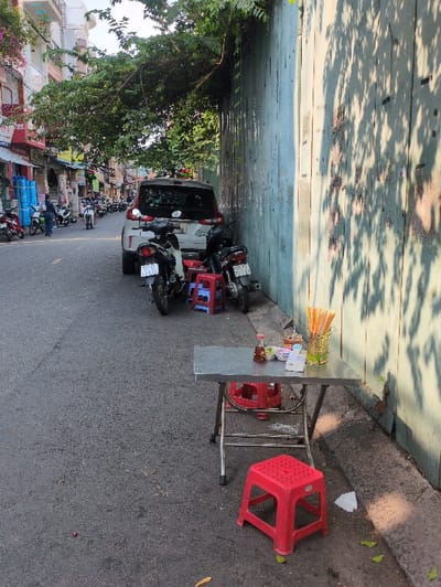 The scene depicts a narrow urban street in Ho Chi Minh City, Vietnam, under bright daylight with dappled sunlight creating strong shadows.
In the immediate foreground, a small, rectangular stainless-steel folding table is positioned on the roadside against a faded light-blue corrugated metal wall. On the table, a clear glass vessel holds numerous yellow chopsticks, alongside a small red-capped condiment bottle, a tissue dispenser, and three small white ceramic dishes. Two red plastic stools are present, one directly in front of the table and another partially visible tucked underneath. On the dark asphalt pavement surrounding the table, a crumpled white tissue, several scattered green leaves, and a small reddish object are visible.
Behind the table, two motorbikes are parked; one is dark-colored and partially obscures a blue plastic stool. Further down the street, a white SUV is parked facing away from the camera, partially on the asphalt and partially near the corrugated wall. A line of additional motorbikes is parked along the left side of the asphalt road.
The left side of the street is lined with multi-story buildings, characteristic of urban Vietnamese architecture, featuring various pastel colors, balconies, and ground-floor storefronts. One shop displays several large, rolled blue items. Several individuals are visible in the mid-distance: one person stands near the blue items, and another is seen riding a motorbike further down the street. A canopy of dense green tree foliage extends over the street from the left, casting shadows onto the corrugated wall. The corrugated wall itself shows signs of wear, including faded paint and darker stains.