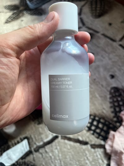 A person is holding a clear plastic bottle of "celimax DUAL BARRIER CREAMY TONER 150 ml / 5.07 fl. oz.". The bottle is half full with a white, opaque liquid. The background is a patterned carpet and a pink fabric. The scene is indoors.