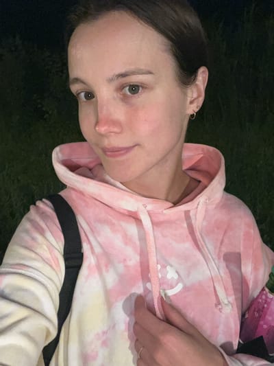 The media file is a close-up, vertical, portrait-style photograph of a young person wearing a pink tie-dye hoodie. The person's face is partially visible, turned to the side, with one eye looking directly at the camera. They have fair skin, light-colored eyes, and dark hair. There is a small hoop earring in their visible ear. The hoodie has a drawstring closure at the neckline. A black strap is visible across the front of the hoodie. The background is dark and appears to be outdoors, possibly at night, with hints of green suggesting grass or foliage. The lighting suggests it is either evening or nighttime. There is no discernible text in the image.
