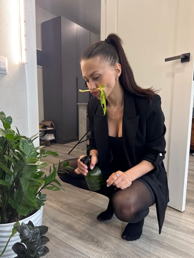 A woman is crouching indoors, attending to potted plants. She has dark hair pulled back, and is wearing a black blazer over a black top, with black tights. She is holding a green spray bottle in her right hand, and there is a green leaf near her face, seemingly held there by her mouth. The plants are in white pots, and the floor is made of light wood. The setting appears to be an apartment or house, with a closed white door with a black handle in the background, and a light fixture and gray cabinets in the background. The photo was taken in Murino, Russia.