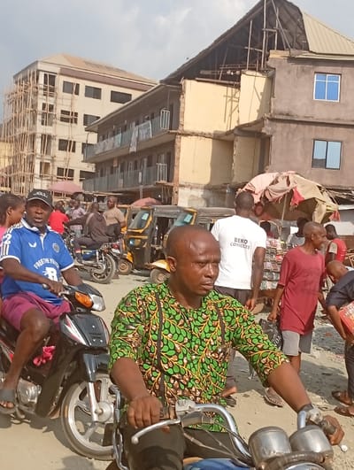 An outdoor shot depicts a busy street scene. Two men on motorcycles are the main subjects, the foreground one wearing a green and yellow patterned shirt. Behind them, another man in a blue jersey rides a motorcycle. In the background, buildings in various states of completion are visible. Pedestrians and auto rickshaws add to the crowded street. The scene captures a lively urban environment under a bright sky.
