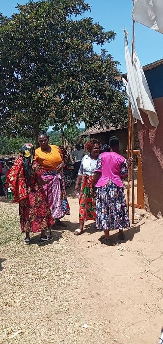This image, rotated clockwise by 90 degrees, presents five women standing outdoors in a sunny, rural setting, likely in Burundu, Kenya. The bright light and distinct shadows indicate it is daytime.
The women are gathered in the mid-ground, each adorned in vibrant, traditional African print attire, often referred to as chitenge or kanga. From left to right, a woman wears an orange top and a multi-colored patterned skirt. Next to her, another woman is dressed in a red and black patterned skirt. Further back, a woman in a white top and a green and red patterned skirt is visible. To her right, a woman in a bright pink top and a blue and white floral patterned skirt is leaning against a wooden structure. Another woman is partially obscured behind this group. Their expressions appear calm, suggesting a casual interaction or gathering.
The setting features a large, lush green tree occupying a significant portion of the left background. To the right, there is a rustic wooden structure, possibly a stall or part of a building, with a white fabric canopy or awning extending from it. The ground is unpaved, consisting of dry dirt and patches of light-colored, dry grass. A dark object, possibly a motorcycle or scooter, is partially visible behind the women in the central part of the image. The clear blue sky is visible above the tree and structures. There is no discernible text in the image.