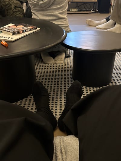 Two round black tables sit on a rug with a black and white dot pattern. A book titled "King's Game" rests on the lower left table. A person in black socks and black clothing sits by the top right table with a light gray garment visible above their sock. Another person with white socks sits across from them in a light gray garment, and a person in camouflage pants sits in the lower left. A television stand with cords is on the left side of the image.