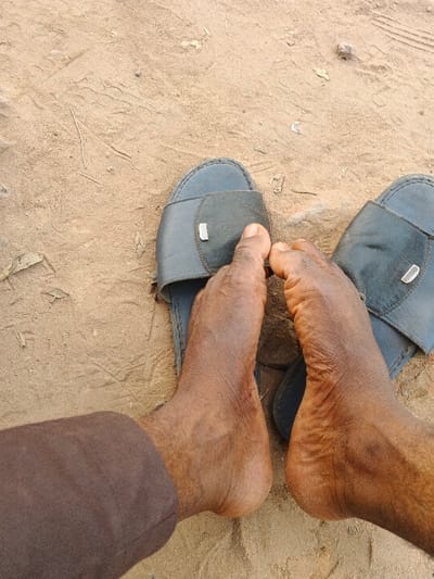A pair of dark-skinned feet are resting on sandals, placed on a sandy surface. The sandals are dark blue, with a simple strap design, each having a small metallic detail. The toes of the feet are visible, extending slightly beyond the edge of the sandals. The skin appears weathered, with visible wrinkles. A portion of a brown fabric can be seen in the lower left corner, suggesting clothing. The ground is dry, with sparse vegetation.
