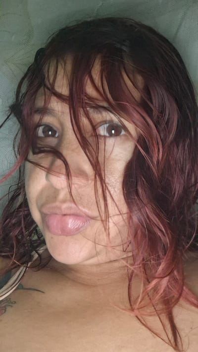 A close-up, waist-up shot of a young adult, likely female, with dark, reddish-brown hair that is wet and styled messily, partially obscuring her face. She is looking directly at the camera with a slight pout. Her skin appears to have a warm tone, and there are faint tattoos visible on her upper chest and shoulder area. The background is a muted, textured green, suggesting an indoor setting or possibly an outdoor area with a softly lit, out-of-focus backdrop. The overall lighting is soft, casting gentle shadows on her face and highlighting the sheen of her wet hair. There is no discernible urban or natural scenery from Vallecito, Venezuela, in this image.
