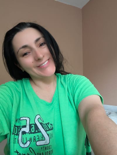 The image shows a selfie of a woman with long, dark hair and a bright green t-shirt. She is smiling warmly, with her head slightly tilted. The background appears to be an indoor setting with neutral-colored walls. The woman's t-shirt has a graphic design on it, featuring large white numbers that resemble "25" outlined with a zig-zag stitching effect. Below this, there is some dark text, partially obscured, with some letters visible such as "T" and what looks like a reversed "J". The overall mood is casual and friendly, suggesting a candid moment captured at home. There are no obvious cues about the time of day or weather. The location context provided is Gelsenkirchen, Germany, though there are no visual elements in the image that specifically indicate this location.