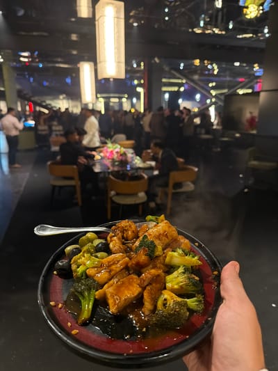A hand holds a plate of food in the foreground, in what appears to be a restaurant or event space. The plate contains pieces of chicken and broccoli in a glossy sauce, with olives scattered around. A fork rests on the edge of the plate. In the background, people are gathered at tables and mingling in a dimly lit environment. The decor suggests a modern establishment with industrial-style lighting fixtures hanging from the ceiling. The scene likely takes place in Gurgaon, India, given the context.