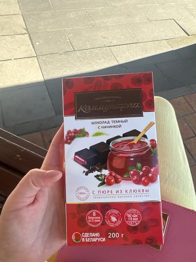 The image shows a person holding a box of dark chocolate with cranberry filling. The box has text in Russian indicating the brand "Kommunarka" and "dark chocolate with filling". There is also an image of chocolate squares, a jar of cranberry puree, and fresh cranberries on the front of the box. The background shows a paved area, possibly a sidewalk, with shadows cast by sunlight. The person's hand is visible on the left side of the image, holding the box. The overall setting appears to be outdoors.