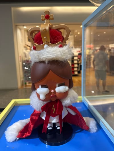 This media file displays a close-up, portrait-oriented photograph of a toy figure. The figure is a cartoon-like representation of a dog, wearing a golden crown adorned with red jewels and a white, furry trim. The crown sits atop the figure's brown head, which also features large white eyes and a small pink nose. The figure is dressed in a red and white outfit with a furry collar and a red sash tied around its neck. The background appears to be inside a display case with a blue base and yellow and white framing. The background visible through the display case is blurred but suggests an indoor retail environment with shelves and products. Reflections in the glass show people and an overhead light. The overall impression is that of a display of merchandise, likely in a store, with the figure being the primary subject. There is no discernible text in the image. The lighting suggests it is daytime, and the setting is likely indoors in a brightly lit area.