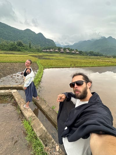Two individuals are captured in a selfie taken in the rice paddies of Mai Chau, Vietnam. The man in the foreground, positioned on the right, wears sunglasses and a black wrap over a white shirt, with his left arm extended towards the camera. To the left, a woman stands on a concrete pathway, looking back at the camera. She has dark hair pulled back and is wearing a black top, a white jacket, and dark blue pants. Behind them, the landscape unfolds with vibrant green and yellow rice fields, reflecting the overcast sky. In the distance, lush, mist-shrouded mountains rise, dotted with traditional stilt houses. The ground appears wet and muddy, indicative of the agricultural setting.