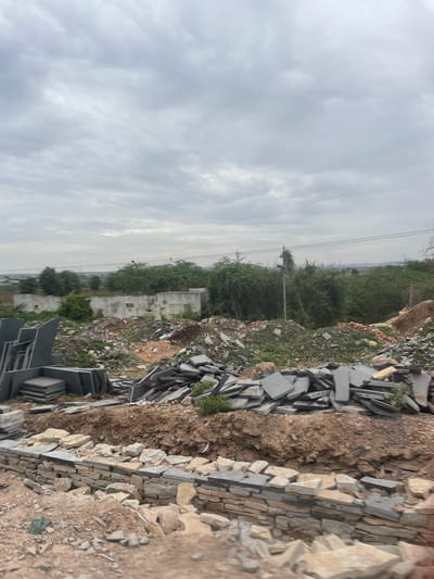 An outdoor, wide-angle, eye-level shot captures a construction or demolition site under a cloudy sky in Bethamcherla, India. In the foreground, a low wall made of rough-hewn stones and flat gray stones is under construction. Piles of various stones, including flat gray slabs and rougher tan-colored rocks, are scattered throughout the scene. To the left, a stack of neatly arranged flat gray stone slabs stands upright. In the midground, a large mound of debris, composed of rubble, dirt, and scattered stones, is visible. Behind the debris, the partially ruined walls of a concrete building are seen, overgrown with vegetation. A line of trees and dense green foliage stretches across the background, with a utility pole and wires interspersed. In the distance, more trees and hints of structures can be seen, with a hazy horizon under the overcast sky. The overall impression is one of an active or recently active site, characterized by raw materials and unfinished work.