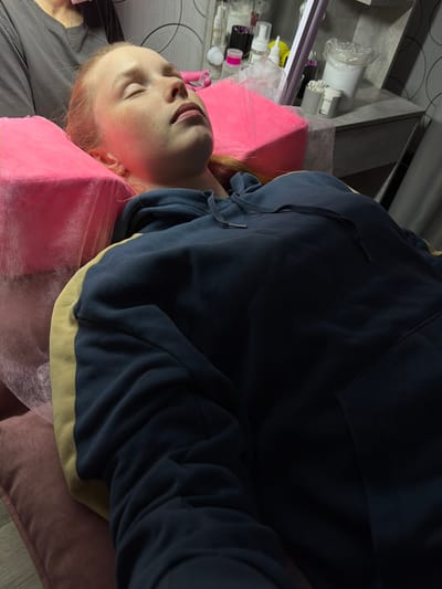 A young person is lying on their back, resting on a pink pillow. They are wearing a dark blue hooded sweatshirt with light yellow trim around the bottom. The person's eyes are closed and their face is visible, revealing light skin and freckles. To the left of the person is a gray or black garment. In the background, there is a shelf holding various cosmetic or personal care products and an obscured gray wall. The scene appears to be indoors, potentially in a salon or spa, but the specific location in the city of Berezniki, Russia cannot be determined based on the image alone.