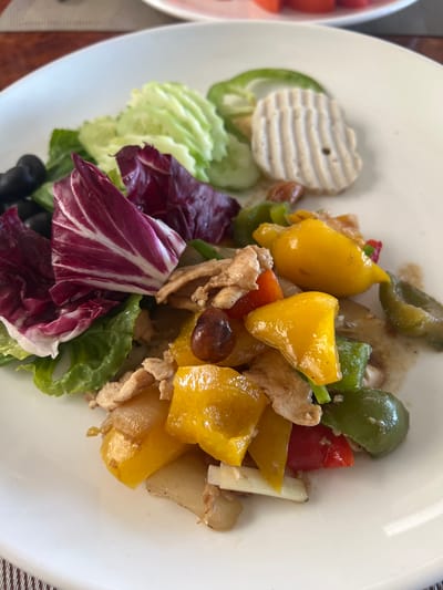 A close-up, eye-level shot captures a white plate of stir-fried chicken and colorful bell peppers, garnished with radicchio, lettuce, olives, and sliced cucumbers. The dish is presented on a brown, woven placemat. The background shows a portion of another white plate with sliced red tomatoes, suggesting a meal setting, possibly in Siem Reap, Cambodia. The lighting is natural, coming from above.