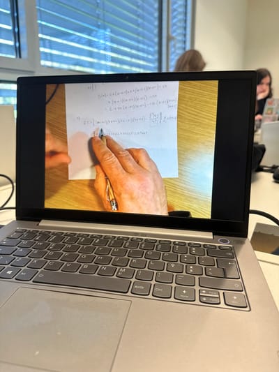 A silver-colored laptop is positioned in the foreground, resting on a white desk surface. Its screen displays a close-up image of a person's left hand, adorned with a ring on the fourth finger, holding a metallic pen and pointing at handwritten mathematical equations on a white sheet of paper. The paper lies on a light brown wooden surface. Visible equations include terms such as `P(u,v)`, `(u-0.1)`, `(0.4-0.1)(0.4-0.2)(0.4-0.3)`, `f'(t)/4T`, and the numerical result `0.7667`. The laptop features a dark grey QWERTY keyboard with a Portuguese layout. A black cable is connected to the right side of the laptop. In the blurred background, a large window fitted with horizontal venetian blinds allows natural light into the room. Two indistinct individuals are visible, seated at desks, suggesting an educational environment such as a classroom or study hall. This scene is situated within a university or educational institution in Guimarães, Portugal.